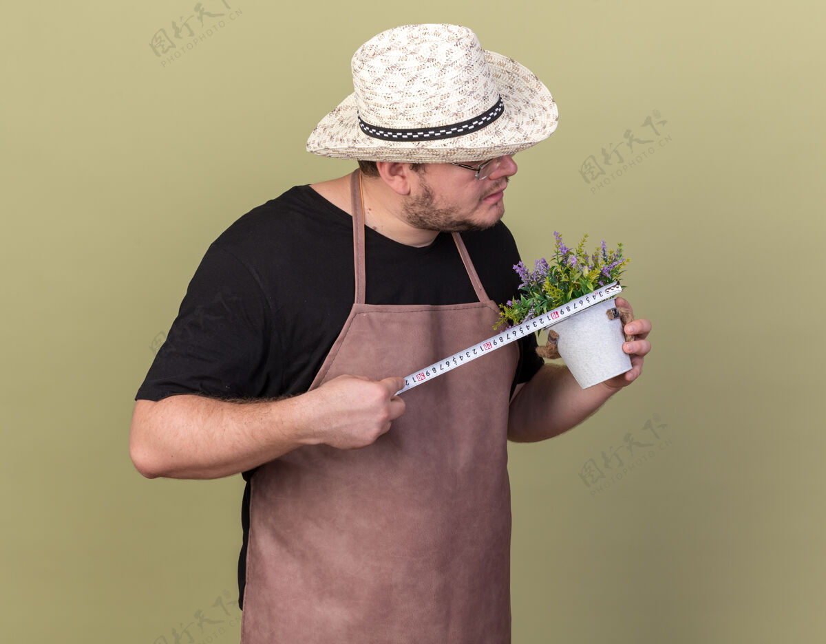 花自信的年轻男园丁戴着园艺帽 用卷尺在橄榄绿的墙上测量花盆里的花朵帽子男性花盆
