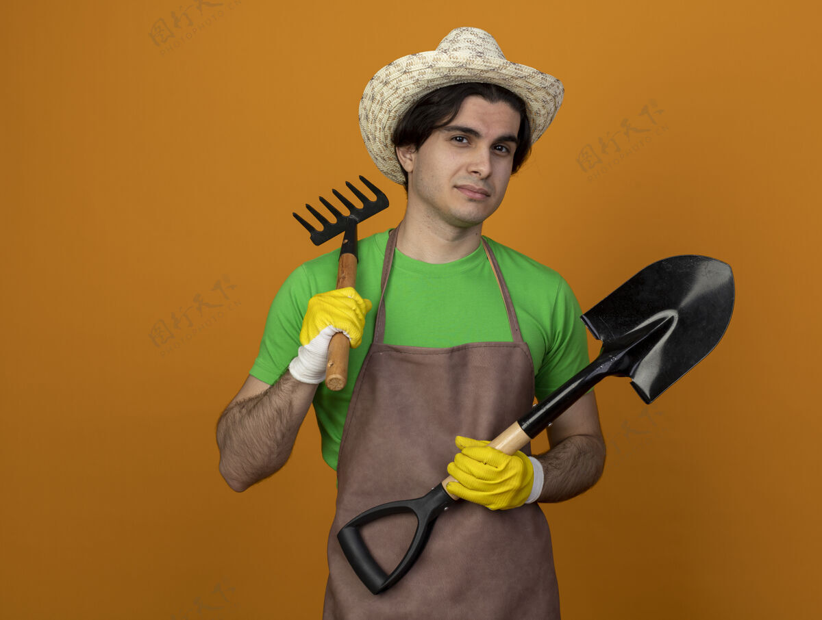 男高兴的年轻男园丁穿着制服 戴着园艺帽 戴着手套 拿着铲子 把耙子放在肩膀上 孤立地放在橘子上 留着复印空间橙色请穿