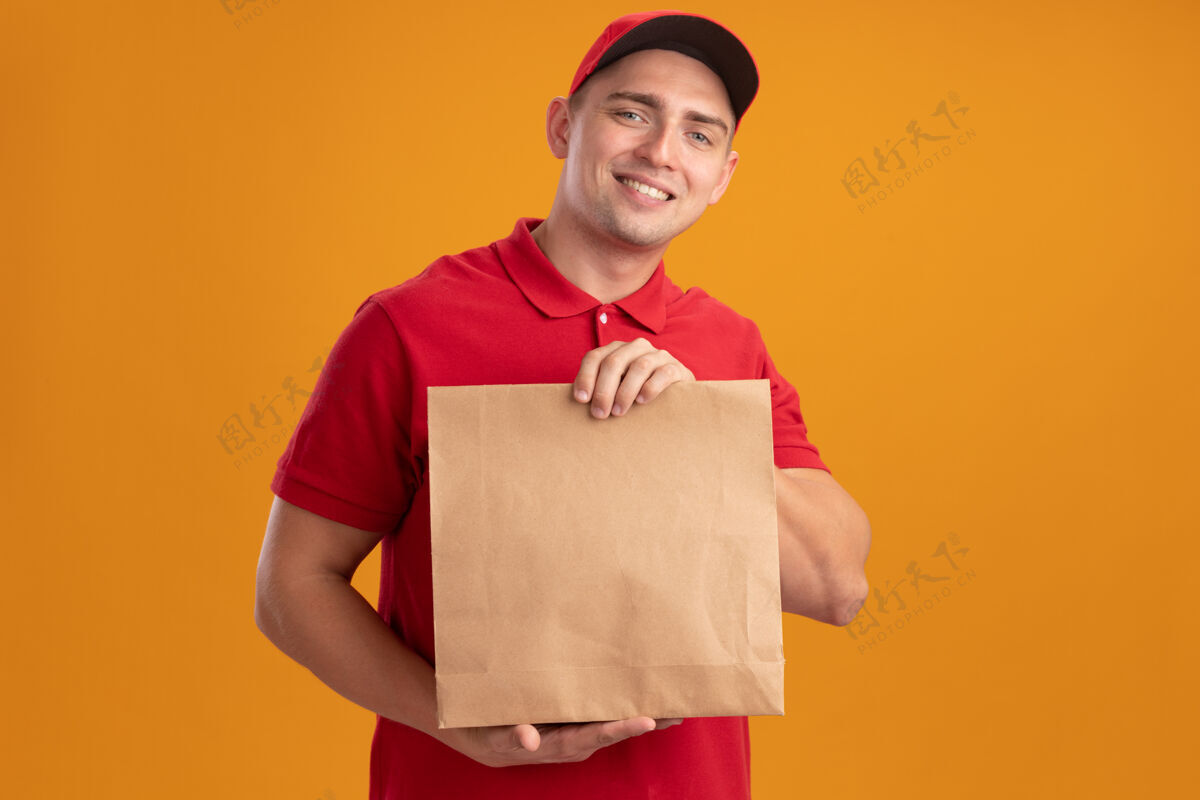帽子微笑着的年轻送货员穿着制服 戴着帽子 手里拿着隔离在橙色墙上的纸食品包送货戴着拿着