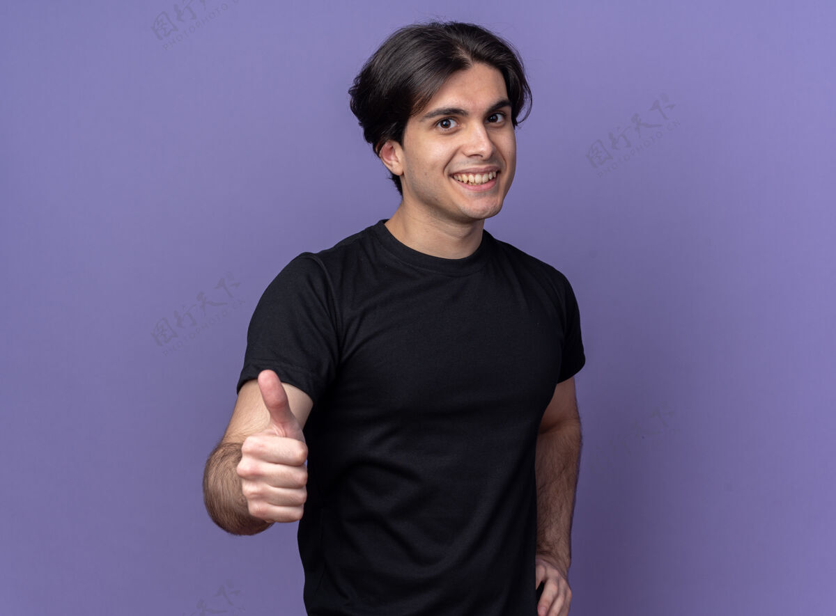 臀部微笑的年轻帅哥穿着黑色t恤 大拇指向上 手放在臀部 紫色的墙上手年轻拇指