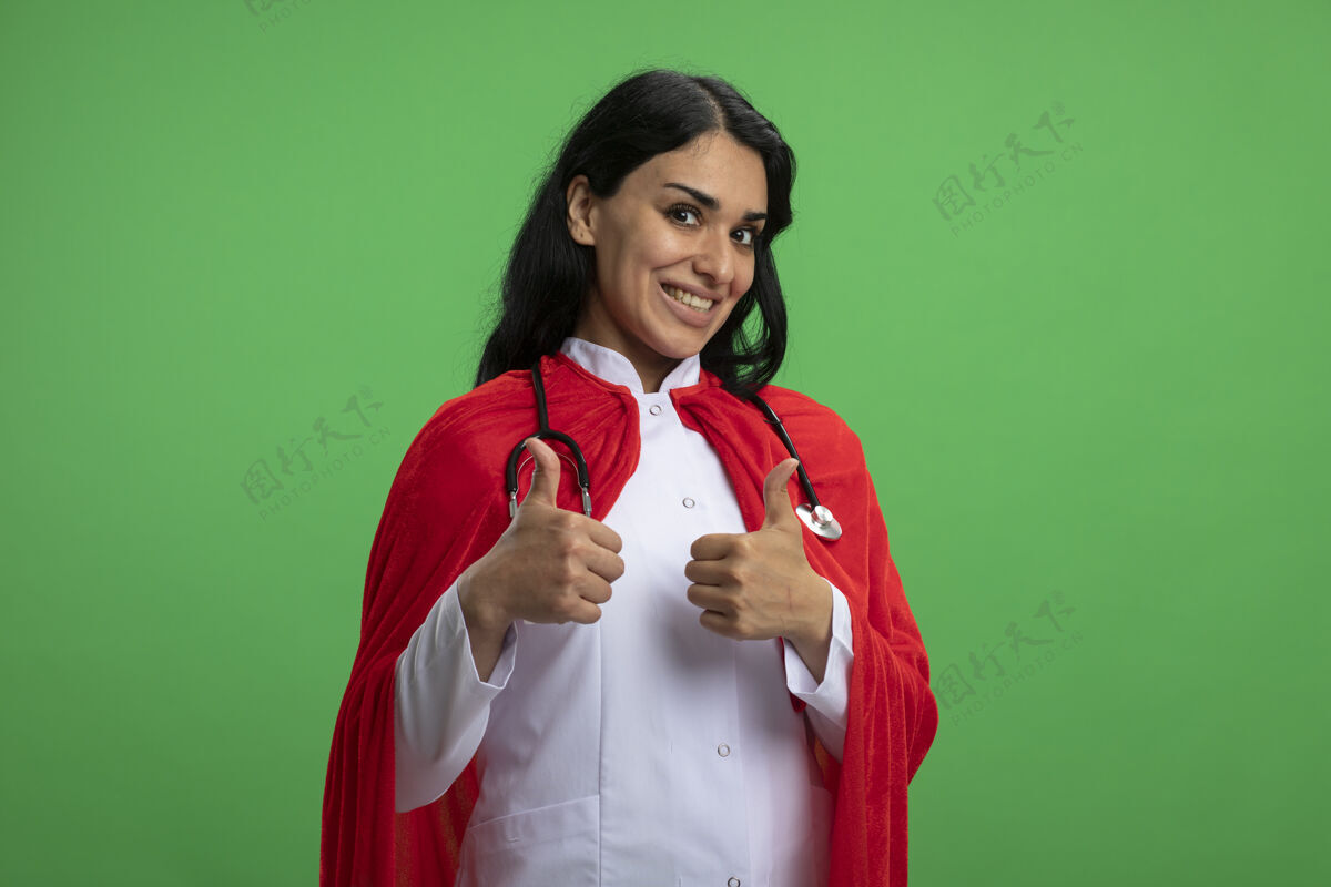 长袍微笑着的年轻超级英雄女孩穿着医用长袍 听诊器在绿色的地面上竖起大拇指年轻拇指医疗