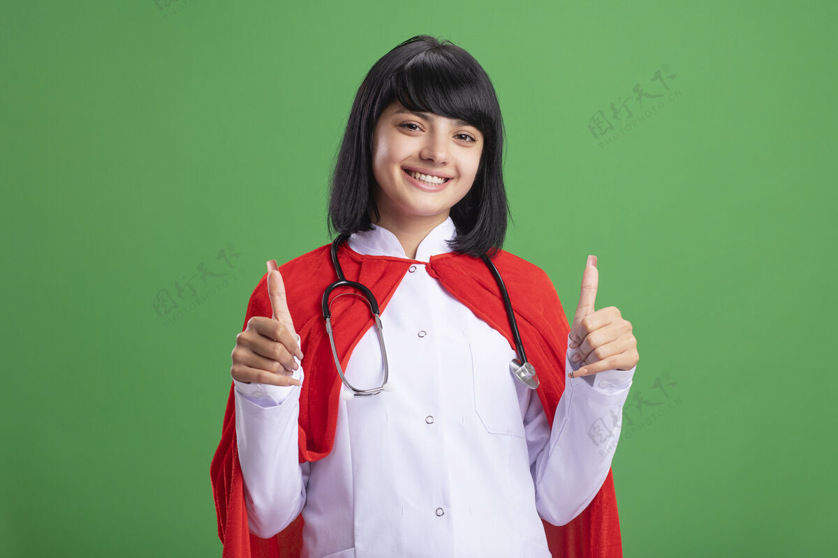 医疗带着微笑的年轻超级英雄女孩戴着听诊器 穿着医用长袍和斗篷 在绿色的草坪上竖起大拇指年轻穿拇指