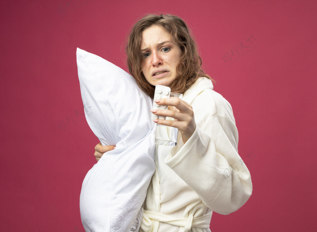 粉红被吓坏了的年轻病女孩穿着白色长袍 抱着枕头 看着一杯水和粉红色的药片隔离开来拥抱枕头水