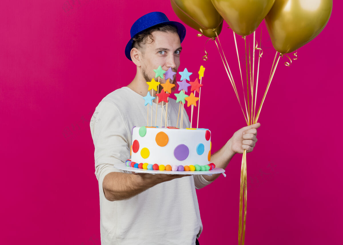 男人高兴的年轻英俊的斯拉夫党家伙戴着党的帽子拿着气球和生日蛋糕与星星伸展向前方看前面的粉红色墙上孤立的复制空间年轻穿着人