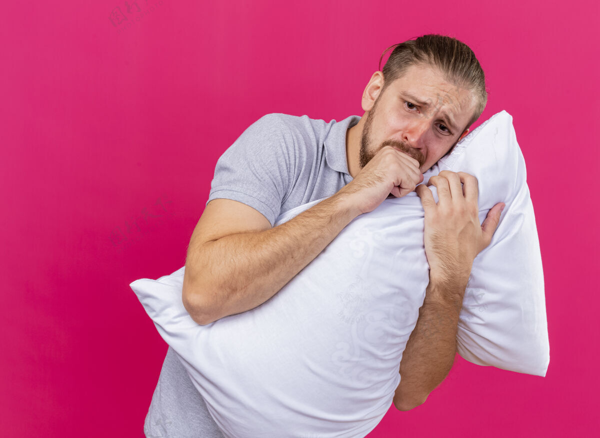 靠近悲伤的年轻英俊的斯拉夫病人抱着枕头看着直咳嗽保持拳头靠近嘴巴隔离在粉红色的墙上复制空间拳头英俊斯拉夫