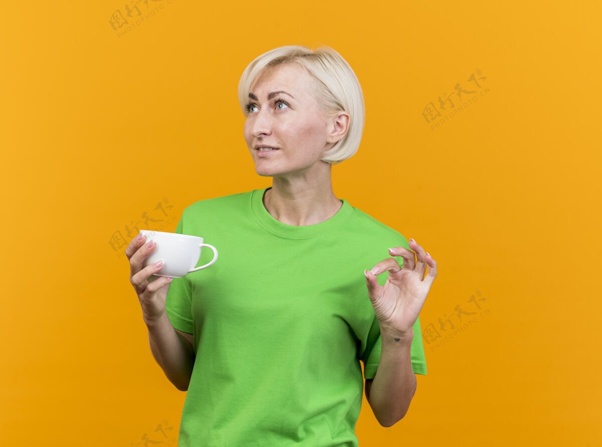 脸高兴的中年金发斯拉夫妇女端着一杯茶看着旁边做ok的标志 隔离在黄色的墙上 留着复制空间人请壁板