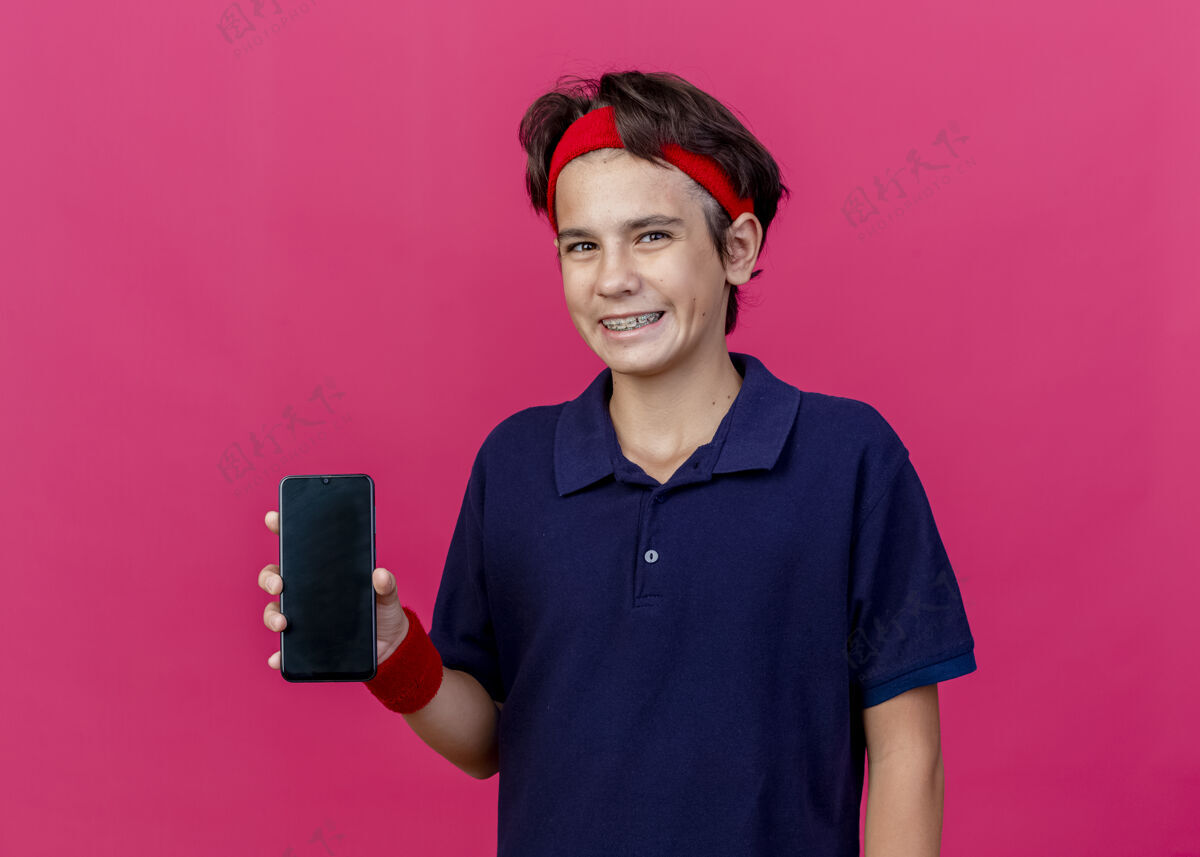 人面带微笑的年轻帅气的运动男孩戴着头带和戴着牙套的腕带 在粉红色的墙上显示手机正看着前面的隔离墙手机微笑电话