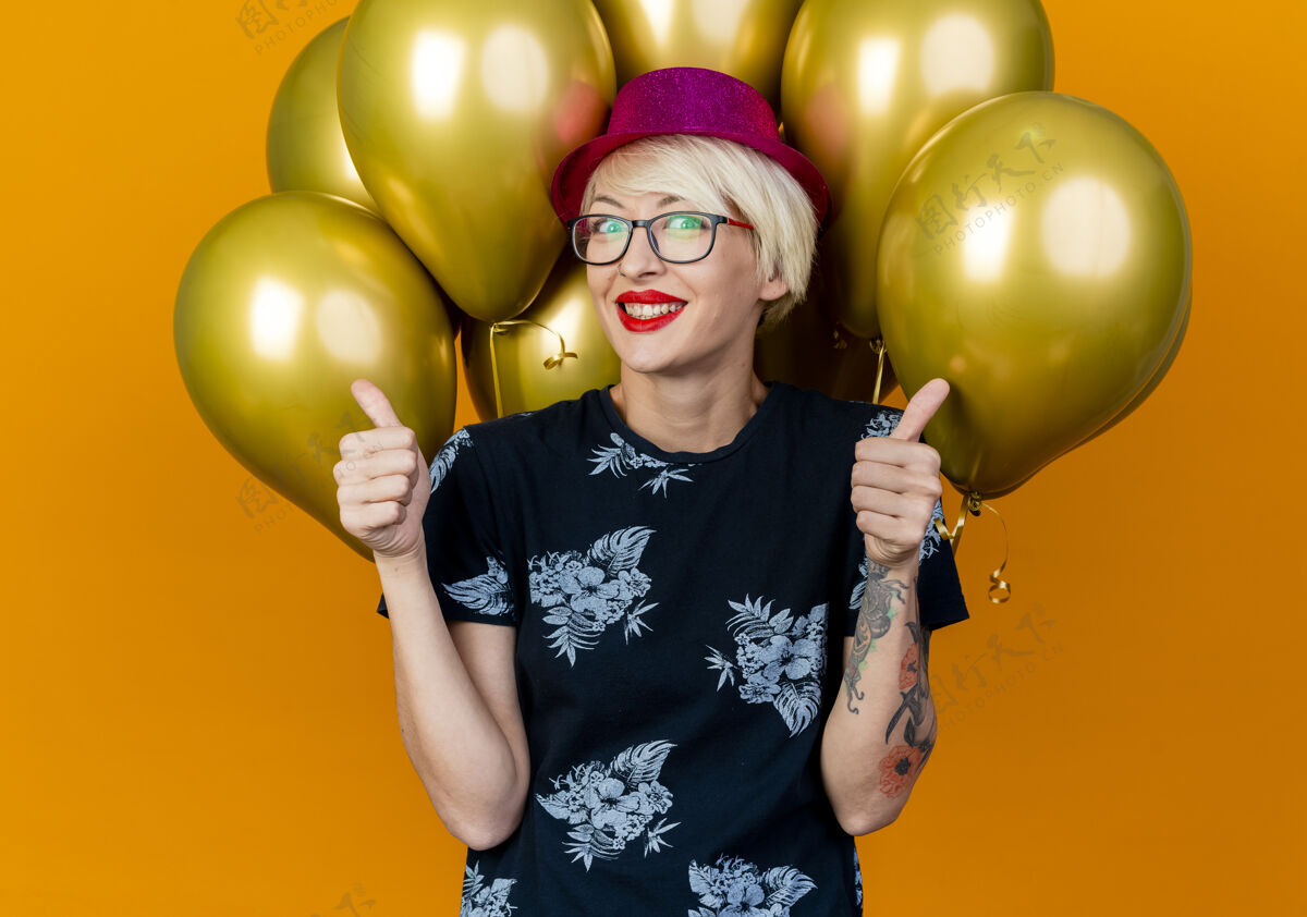 人微笑着的金发派对女郎戴着派对帽戴着眼镜站在气球前看着前面竖起大拇指孤立地站在橙色的墙上衣服穿着眼镜