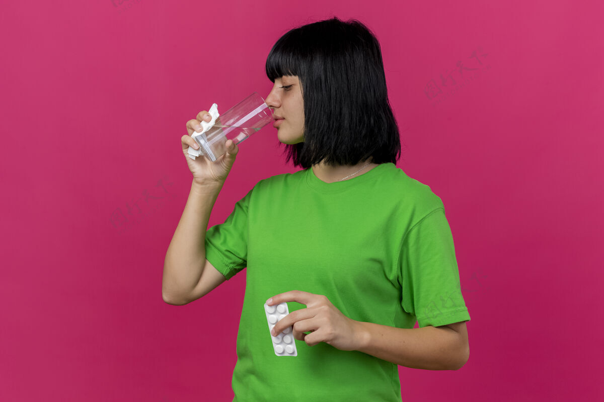 感情年轻生病的女人拿着一包药片 餐巾纸和一杯水 饮用水 隔离在粉红色的墙上 留着复印空间人疾病衣服