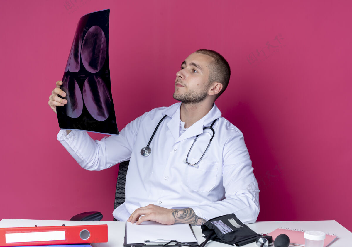 X光年轻的男医生穿着医用长袍和听诊器坐在办公桌旁 手里拿着工作工具 看着隔离在粉色背景上的x光照片射击手持听诊器