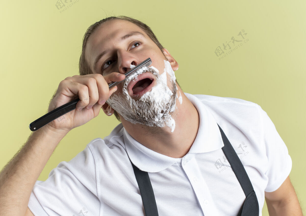 直年轻帅气的理发师穿着制服刮胡子用直剃刀看着脸上涂着剃须膏隔离在橄榄绿的背景上橄榄制服帅气