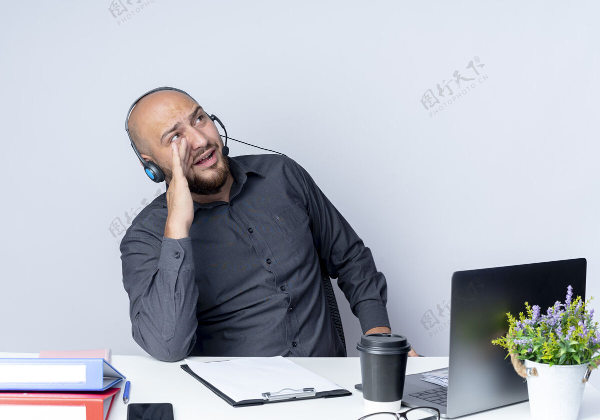 电话年轻秃顶的呼叫中心男子戴着耳机坐在办公桌旁 手里拿着工作工具 抬头对着一个被隔离在白色背景下的人喊叫中心工作坐着