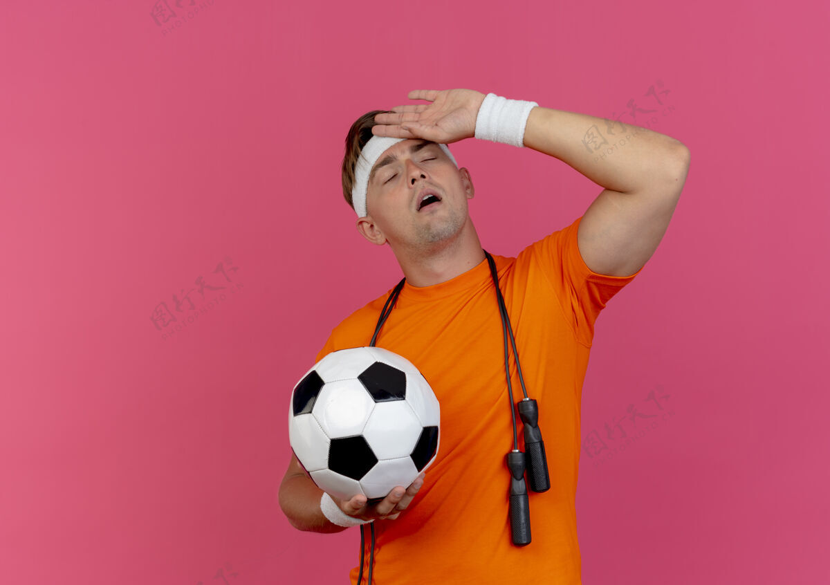 腕带疲惫的年轻英俊的运动型男子戴着头带和腕带 脖子上系着跳绳 手里拿着足球 手放在额头上 闭着眼睛 隔离在粉色背景上 有复制空间眼睛年轻围着