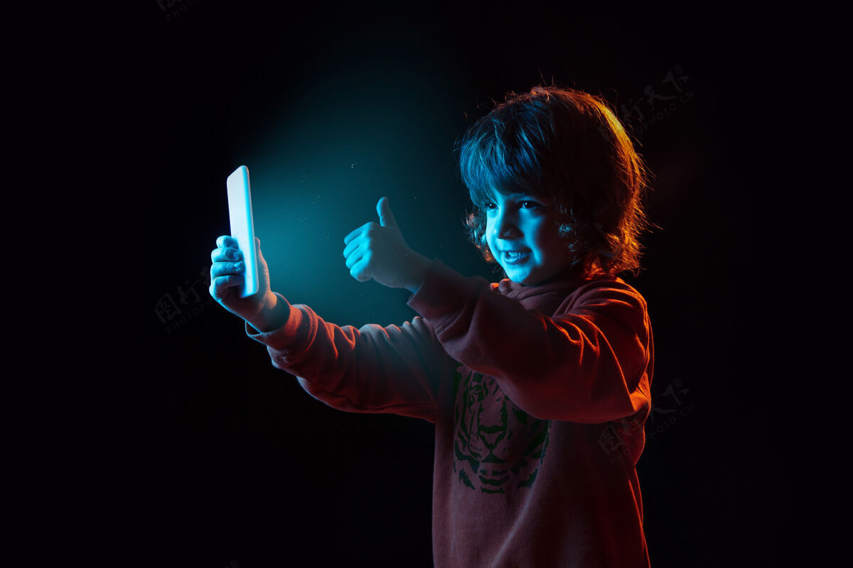 视频日志用智能手机拍照 竖起大拇指霓虹灯下深色背景上的白人男孩肖像漂亮的卷曲模型人类情感的概念 面部表情 销售 广告 现代科技 小玩意在线表情公司