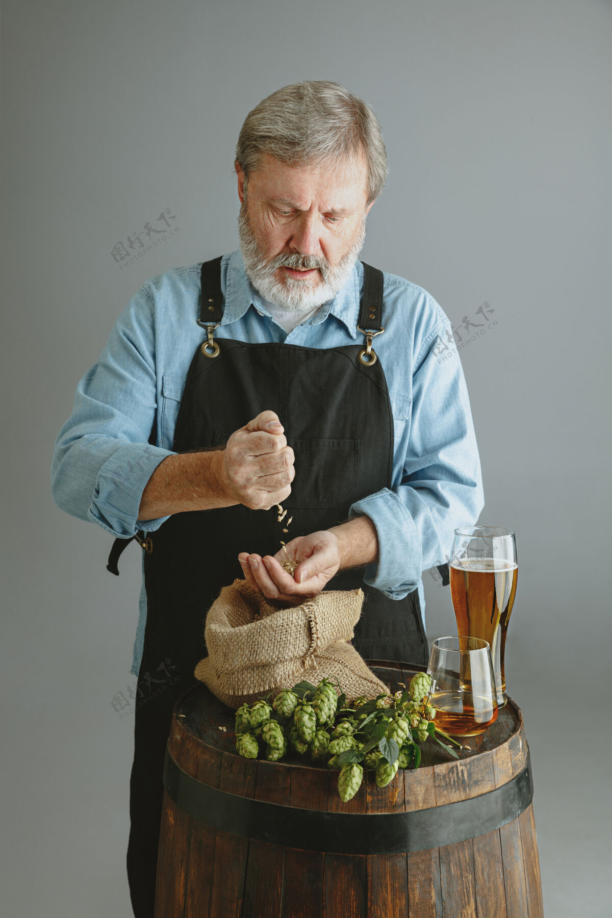 酿酒自信的高级酿酒师 在灰色墙壁上的木桶上 用自制的玻璃啤酒工厂老板展示了他的产品 检验质量酒吧微型围裙