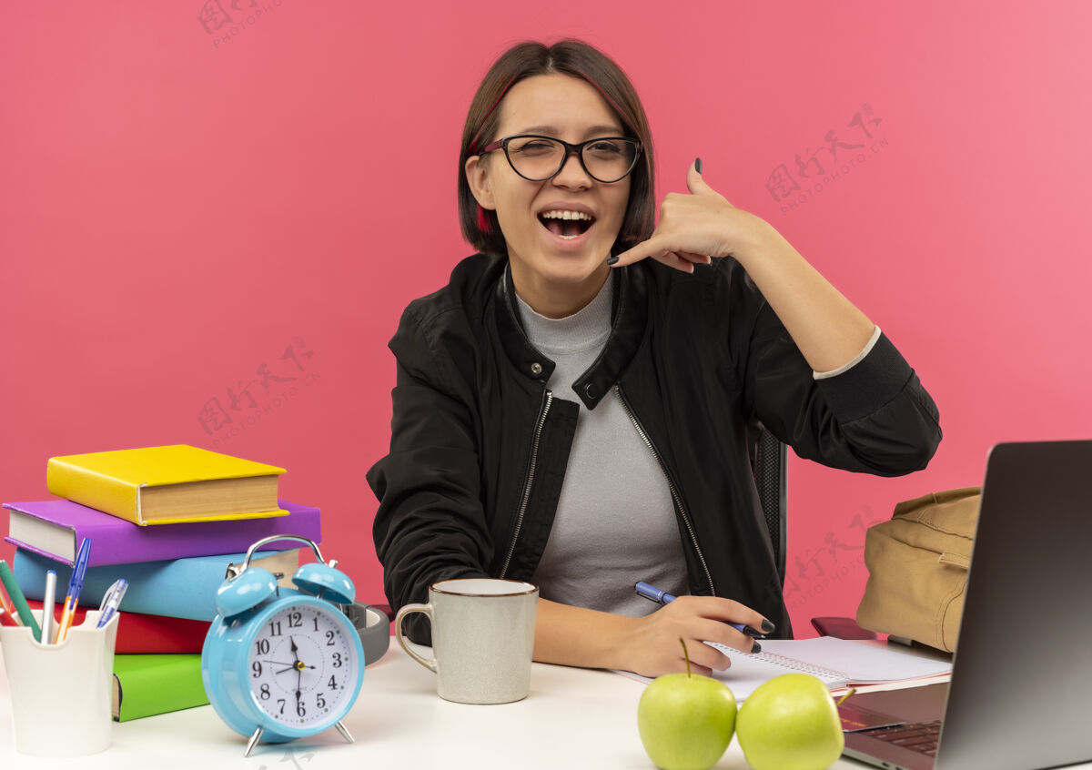 快乐快乐的年轻女孩戴着眼镜坐在书桌旁做打电话的手势做作业孤立的粉红色学生粉色电话