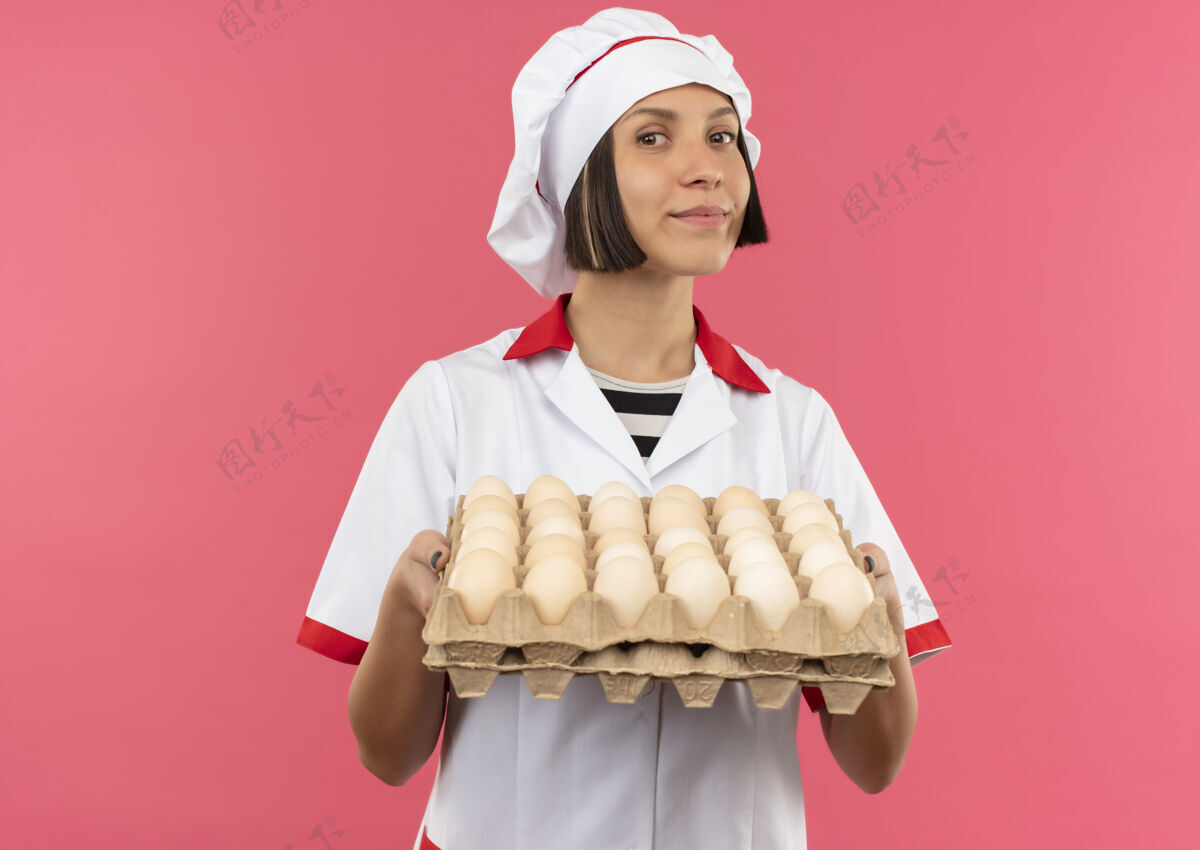 纸箱身着厨师制服的年轻女厨师手拿一盒鸡蛋 鸡蛋被隔离在粉色背景上 并留有复印空间制服厨师鸡蛋