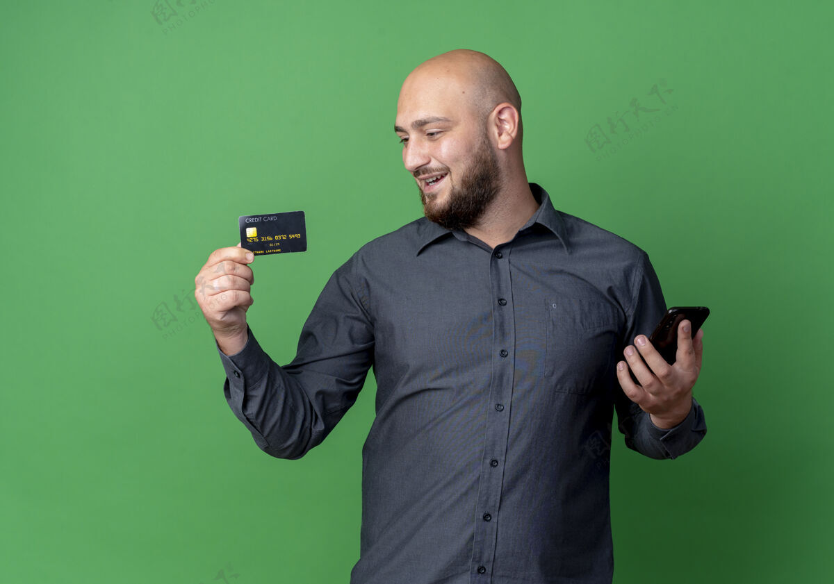 信用快乐的年轻秃头呼叫中心男子手持信用卡和手机看着卡上的绿色隔离男人年轻秃头