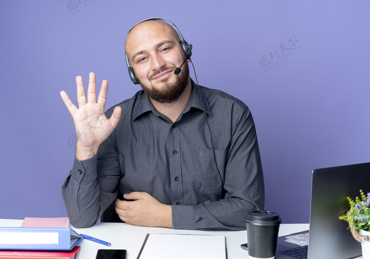 相机高兴的年轻秃头呼叫中心男子戴着耳机坐在办公桌旁的工作工具 向隔离在紫色背景下的摄像头打招呼年轻请坐着
