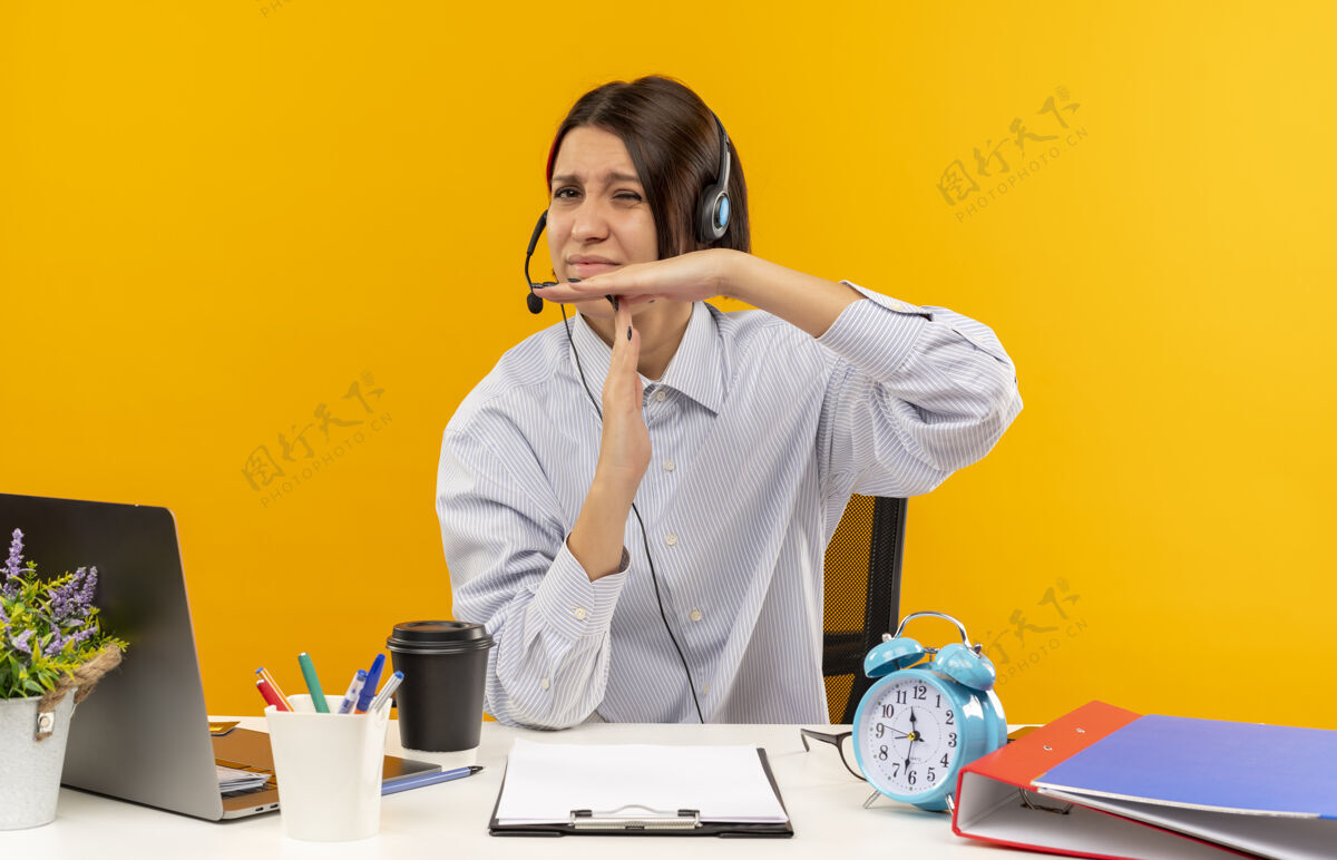 超时未出租的年轻呼叫中心女孩戴着耳机坐在办公桌旁 与工作工具做超时手势隔离在橙色背景上女孩年轻坐着