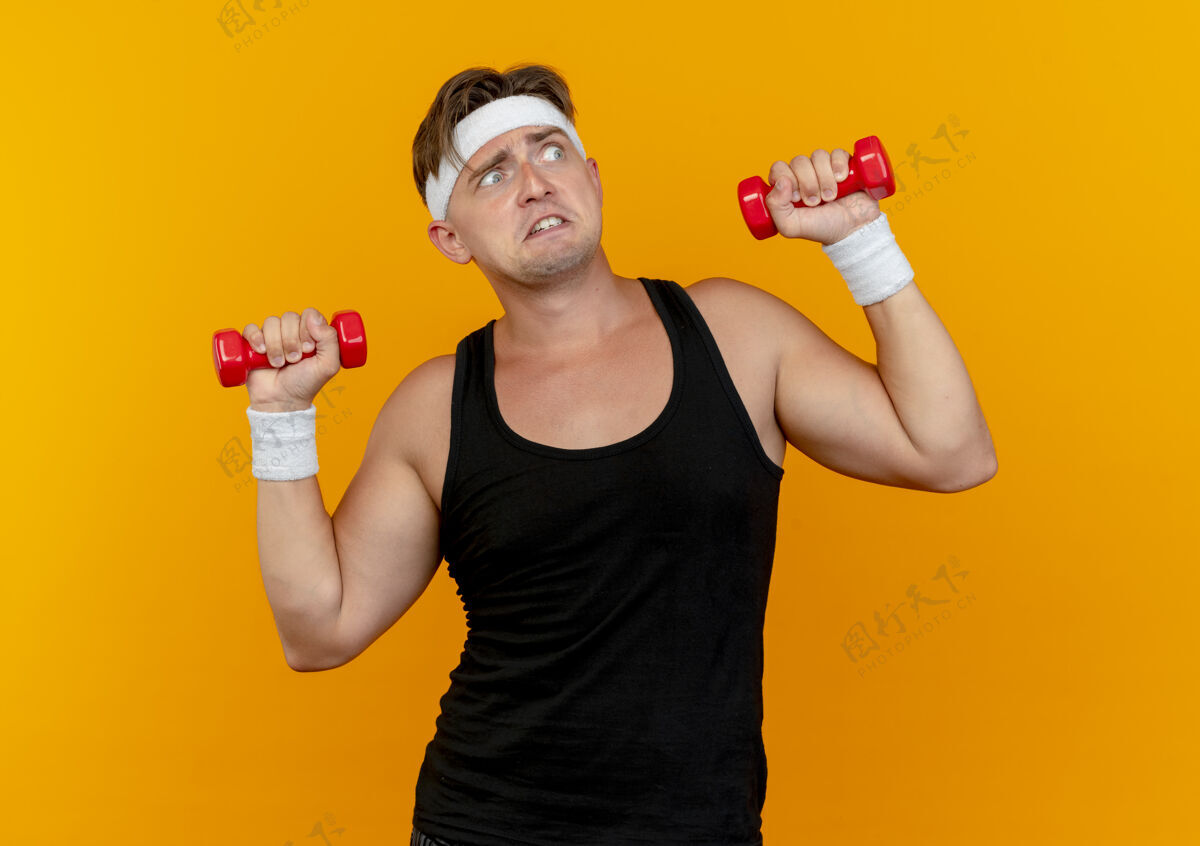 男人印象深刻的年轻英俊的运动男子戴着头带和腕带举着哑铃看着边上孤立的橙色腕带提高运动