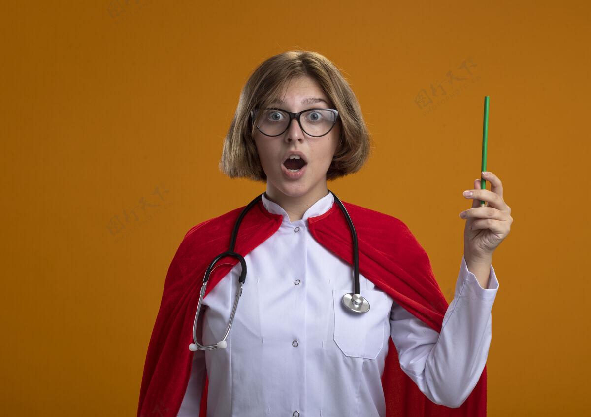 铅笔令人印象深刻的年轻金发女超级英雄在红色斗篷穿着医生制服和眼镜听诊器拿着铅笔看前面孤立的橙色墙壁与复制空间制服金发眼镜