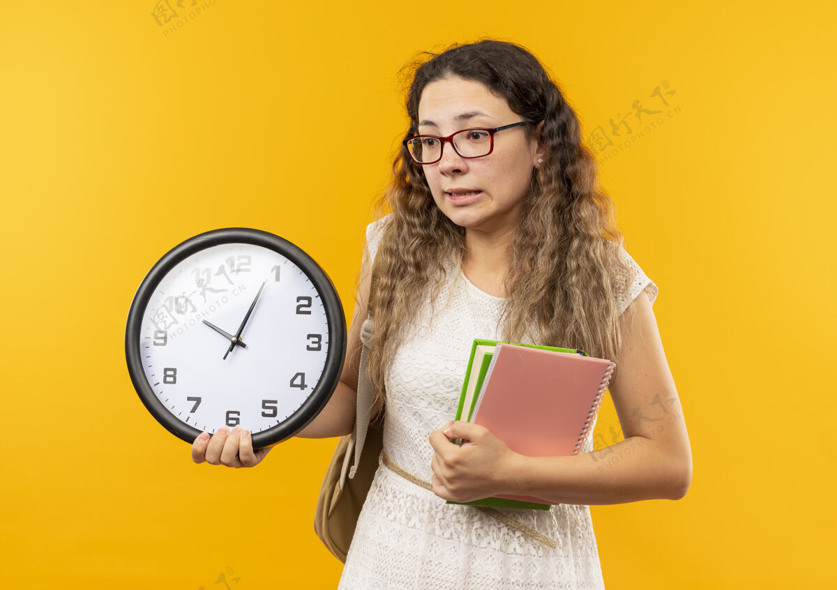 纸条印象深刻的年轻漂亮的女学生戴着眼镜 背着书包拿着时钟 笔记本 看着边上孤立的黄色抱穿黄色