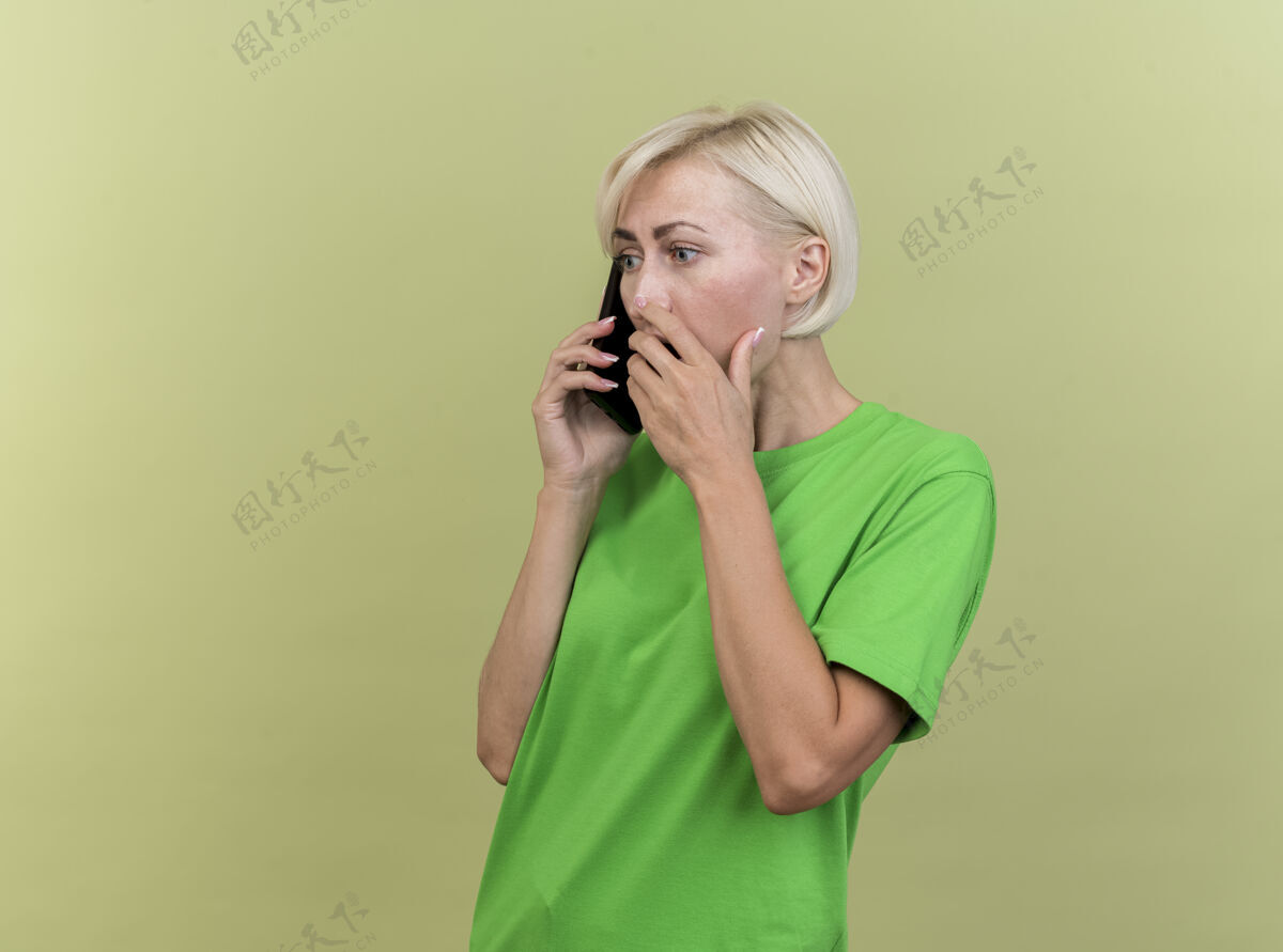 脸惊讶的中年金发斯拉夫妇女站在侧视图讲电话保持手放在嘴上橄榄绿的墙壁与复制空间隔离手斯拉夫轮廓