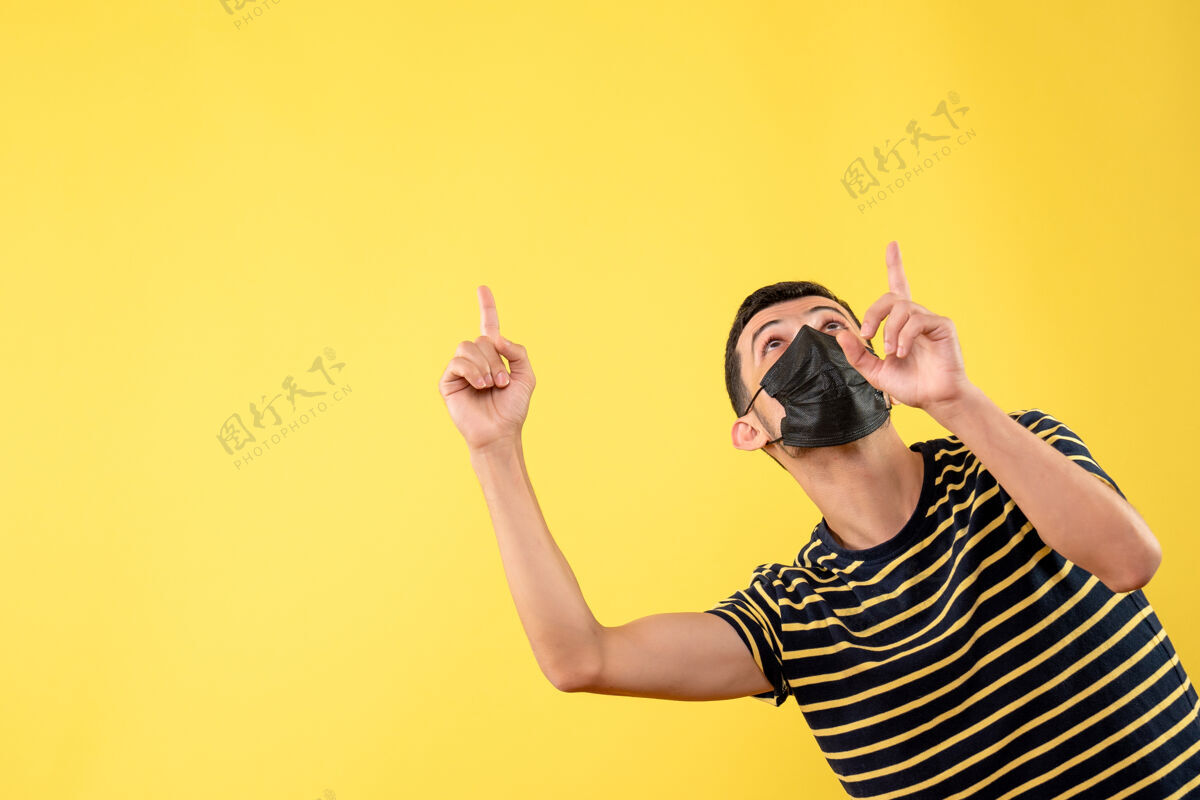 天花板正面图：戴着黑色面具的帅哥指着黄色隔离背景上的天花板成人指向面具
