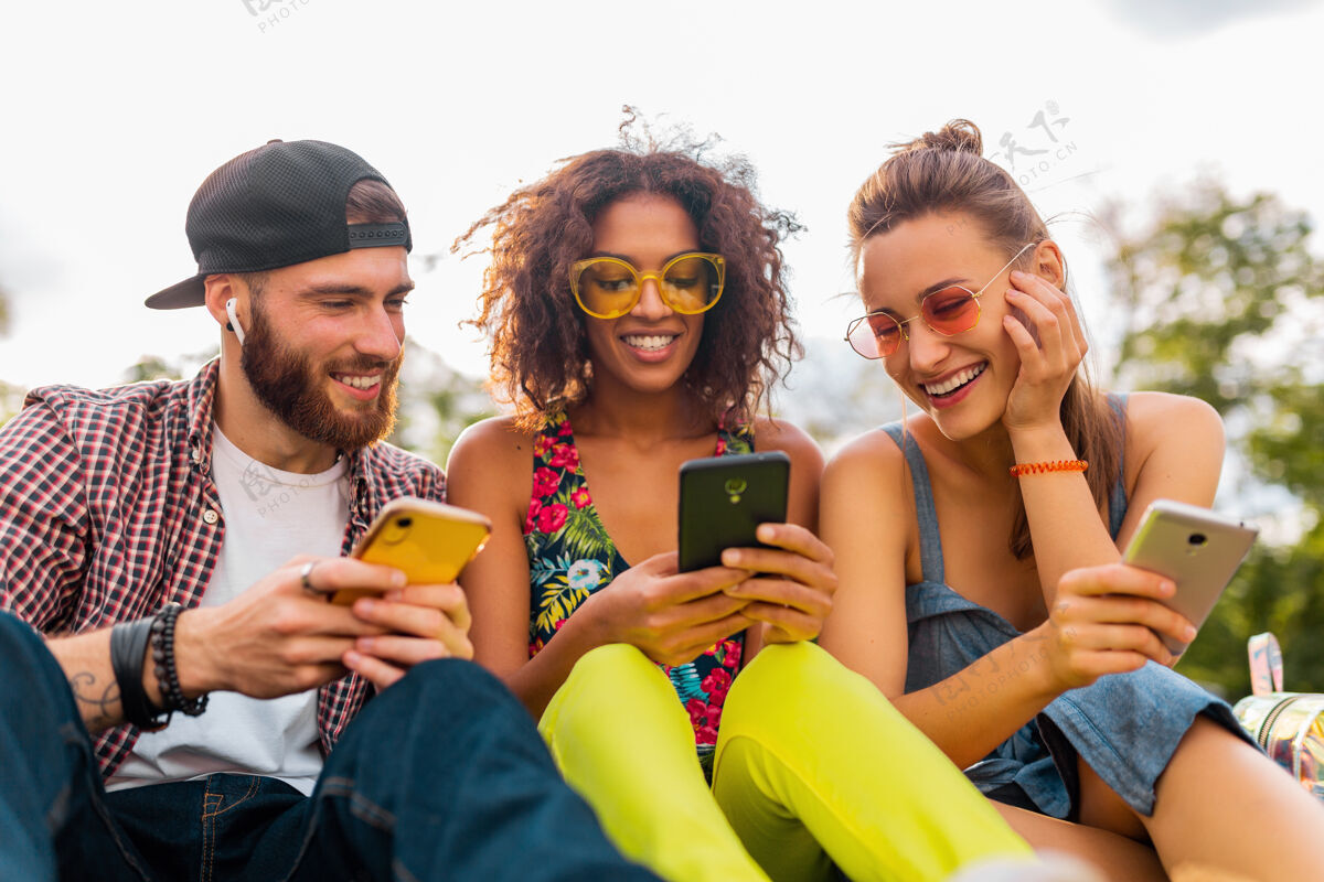 人快乐的年轻朋友们坐在公园里用智能手机微笑 男人和女人一起玩电话多民族积极