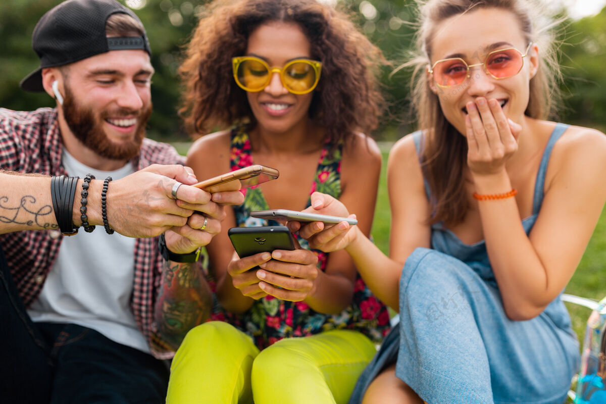 微笑快乐的年轻朋友们坐在公园里用智能手机微笑 男人和女人一起玩团体多种族耳机