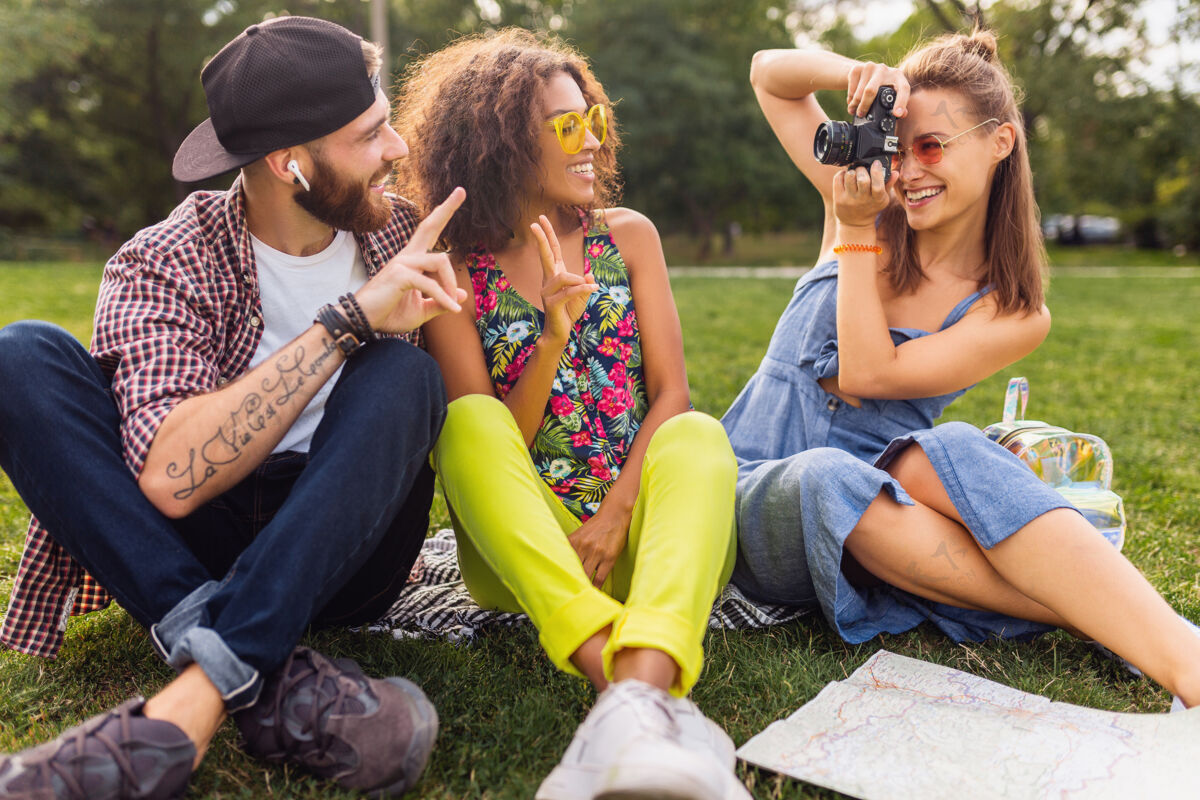 旅行快乐的年轻朋友陪伴坐在公园里 男女同乐 缤纷的夏日潮人时尚风格 带着相机旅行 拍照留念多种族女人女孩