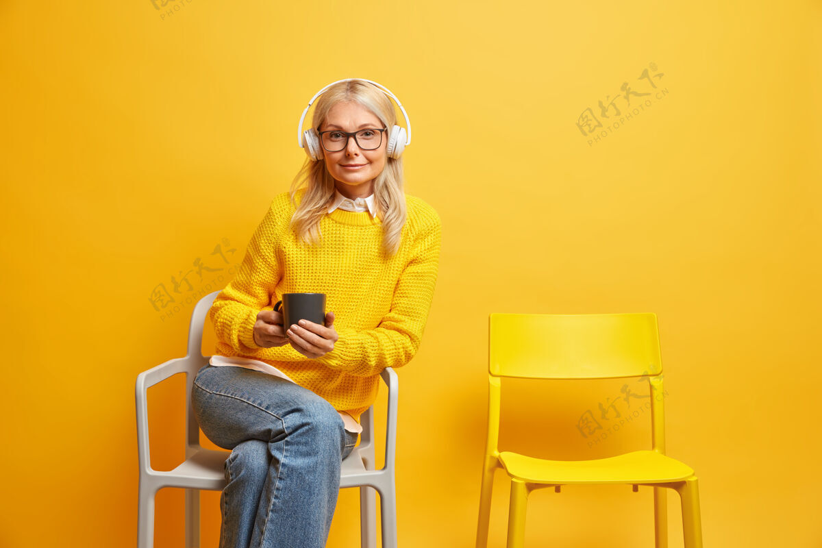 白种人中年妇女坐在舒适的椅子上戴着透明眼镜端着咖啡杯通过耳机听音乐年龄举行金发