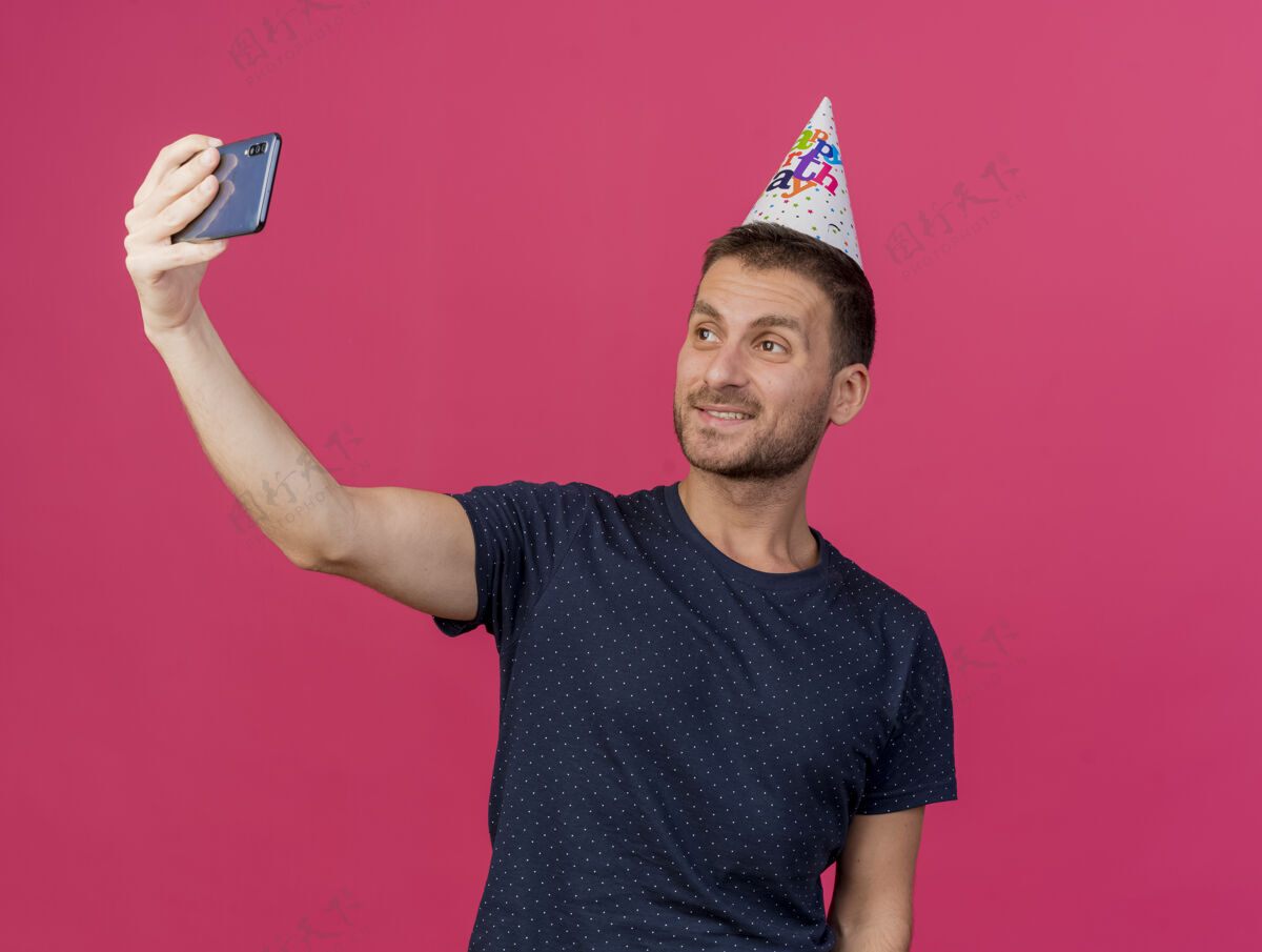 复制微笑英俊的白人男子戴着生日帽举行 并在手机上看自拍的粉红色背景与复制空间隔离生日电话粉色