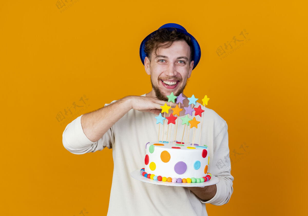 手快乐的年轻英俊的斯拉夫人聚会的家伙戴着聚会帽拿着生日蛋糕和星星看着相机保持手在下巴下隔离在橙色背景与复制空间年轻人星星复制
