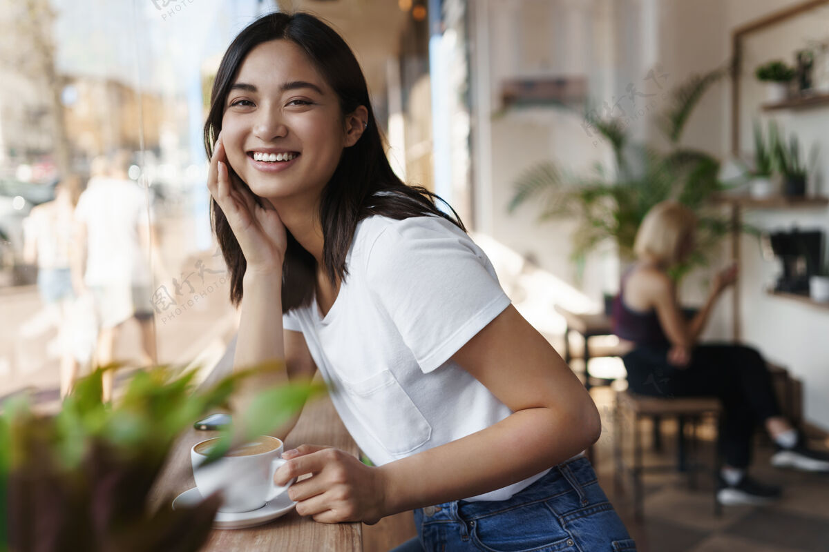 卡布奇诺快乐的亚洲女人坐在靠近窗户的餐厅里对着镜头微笑桌子咖啡馆年轻人