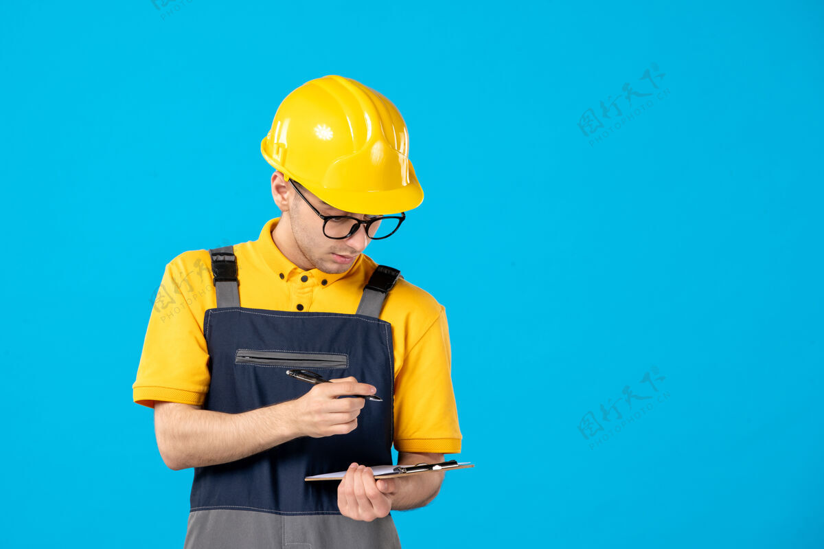 蓝色身穿制服和头盔的男建筑工人在蓝色墙上做笔记的正面图修理工制服工人