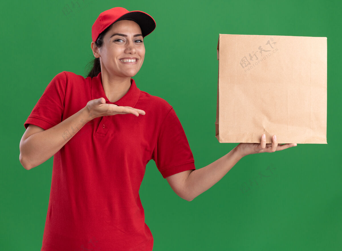 点微笑着的年轻送货女孩穿着制服 戴着帽子 指着隔离在绿色墙上的纸食品包装年轻帽子包装