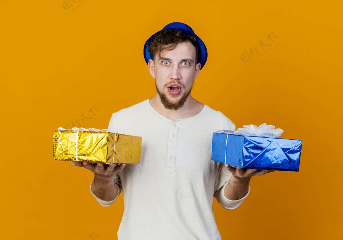 礼物惊讶的年轻英俊的斯拉夫党人戴着党的帽子拿着礼品盒看着相机孤立的橙色背景抱着男人年轻人