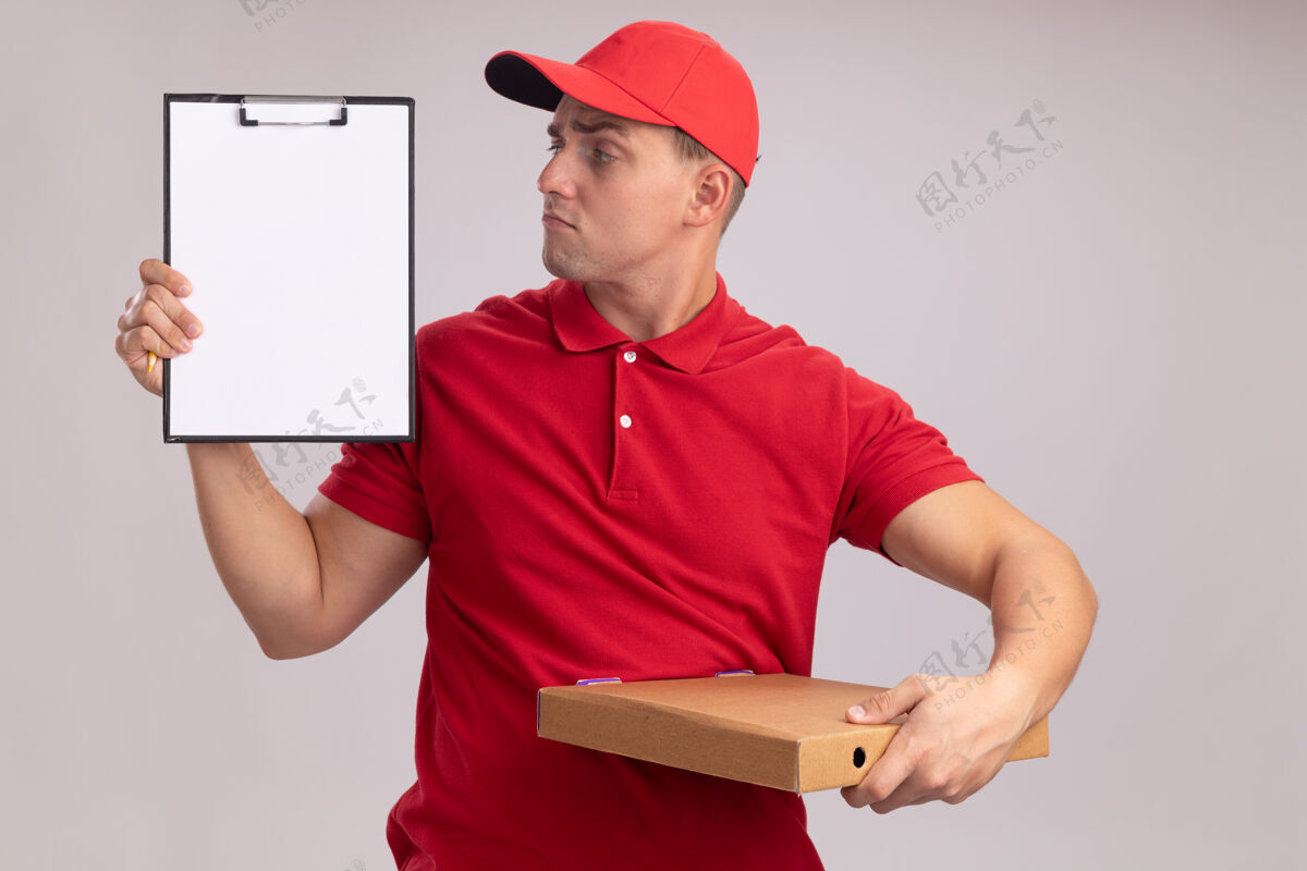 年轻人令人印象深刻的年轻送货员穿着制服 戴着帽子 拿着披萨盒 看着他手上的剪贴板孤立在白墙上制服剪贴板盒子