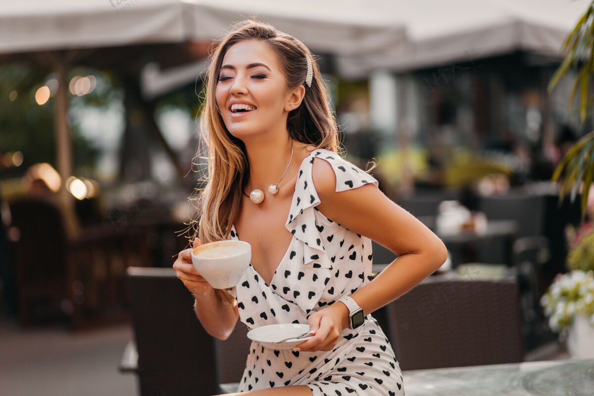 乐趣美丽的微笑的女人穿着时尚的白色印花连衣裙坐在街上的咖啡馆里喝着一杯卡布奇诺女士咖啡馆夏天
