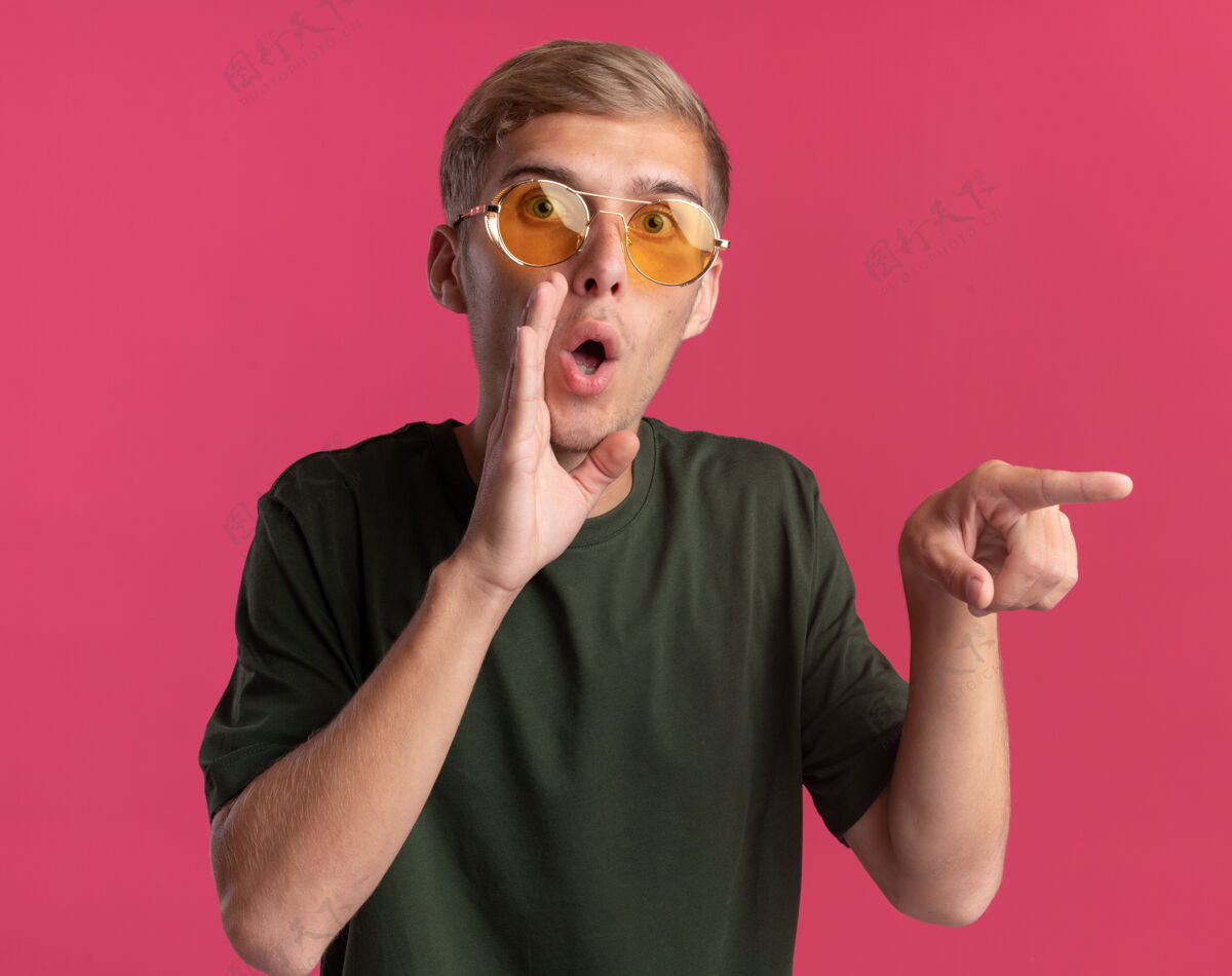 穿可疑的年轻帅哥穿着绿色的衬衫 戴着眼镜 在粉色的墙上低声地指着隔离的一侧年轻可疑点