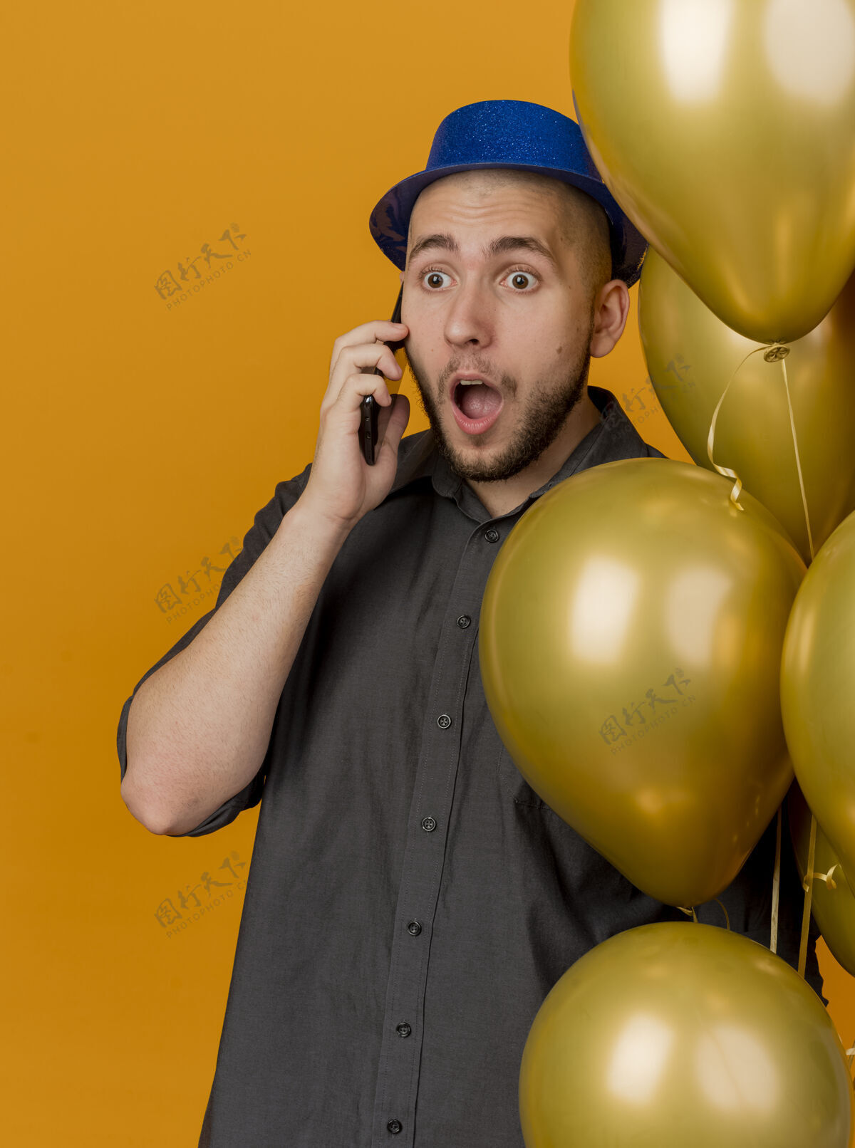 气球一个年轻英俊的斯拉夫人戴着派对帽拿着气球在电话里说话 在橙色的背景下显得很孤立惊喜抱着年轻人