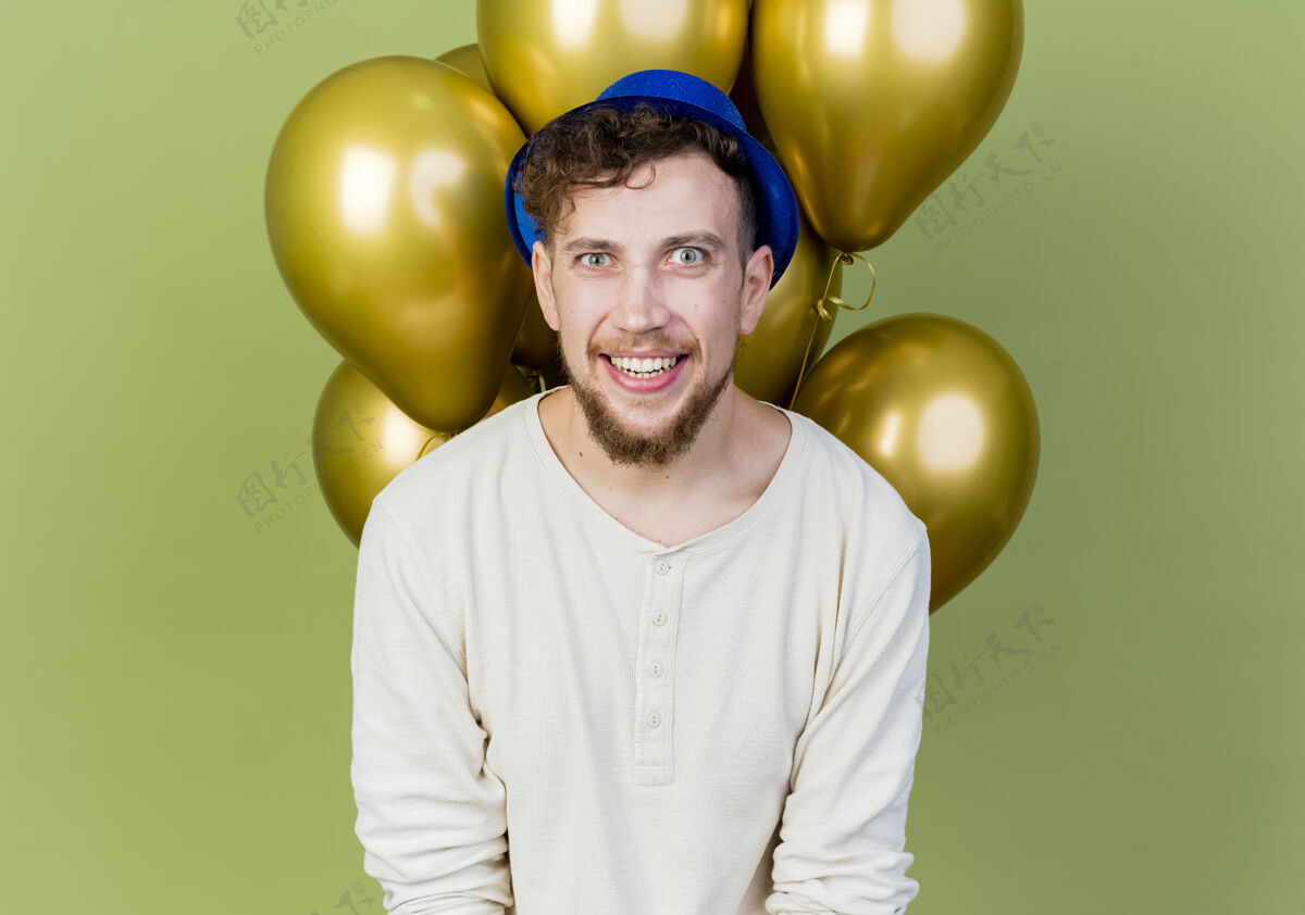 站在前面面带微笑的年轻英俊的斯拉夫党人戴着党的帽子站在气球前看着橄榄绿背景上孤立的相机微笑绿色派对