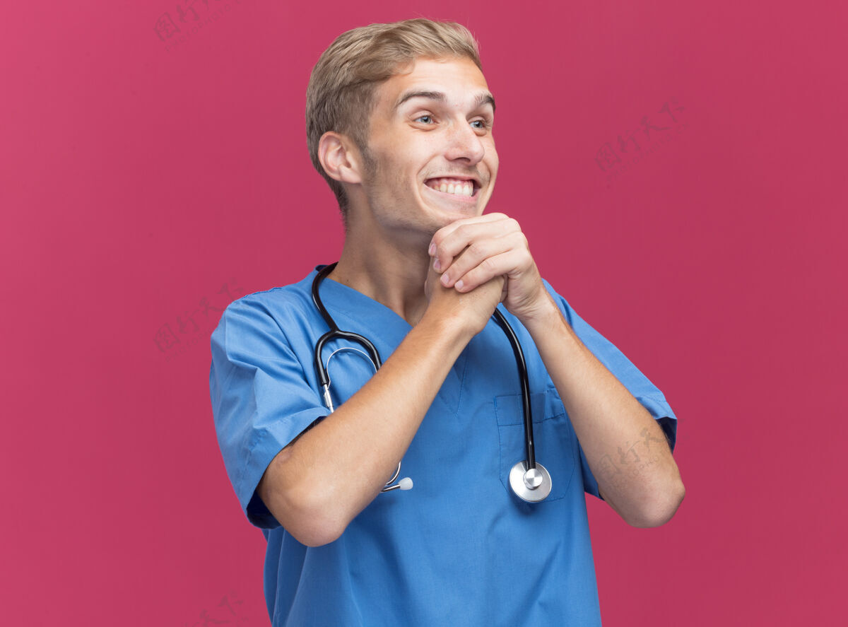 侧身微笑着看着身边年轻的男医生穿着医生制服手持听诊器手托下巴隔离在粉红色的墙上男性下巴手持