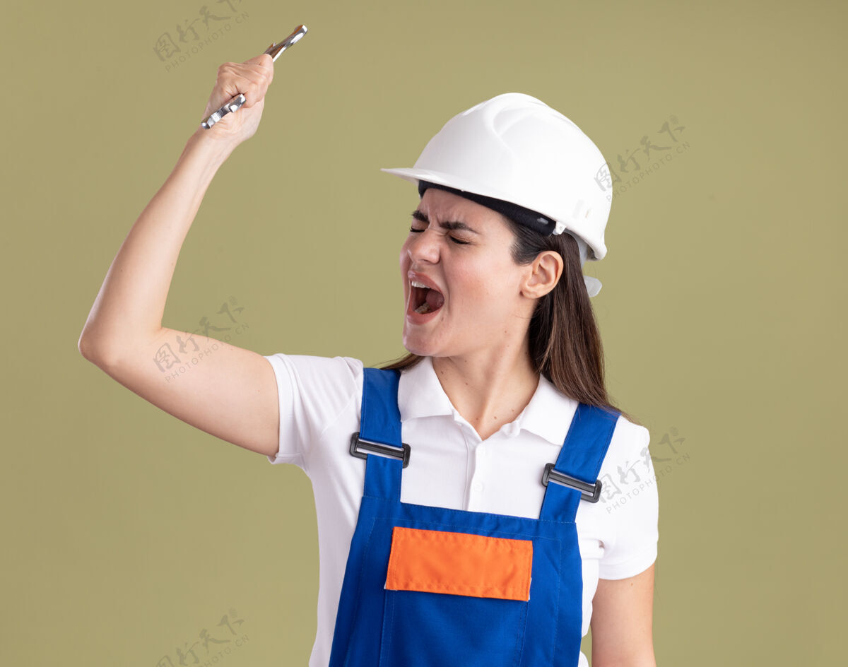 年轻愤怒地闭上眼睛年轻的建筑工人妇女在统一举行开放式扳手隔离在橄榄绿的墙上建筑工关闭举行