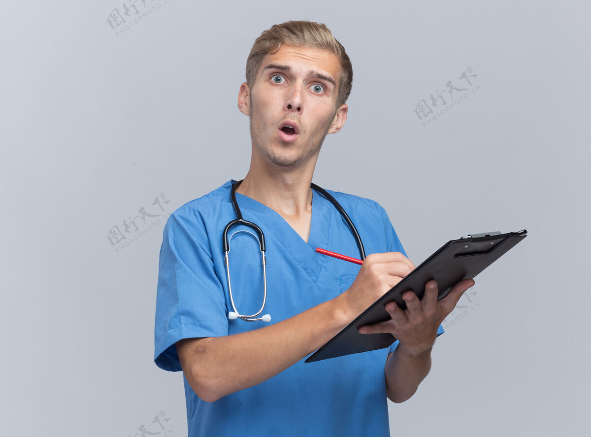 穿什么年轻的男医生穿着医生制服 带着听诊器 惊讶地在白墙上的剪贴板上写着什么医生惊喜年轻人