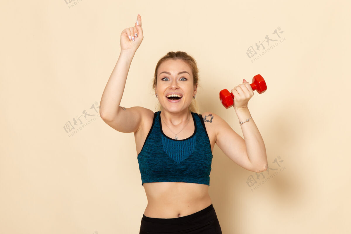 视野正面图身着运动装的年轻女性手持红色哑铃在浅白的墙上运动身体健康美容锻炼健身轻年轻女性