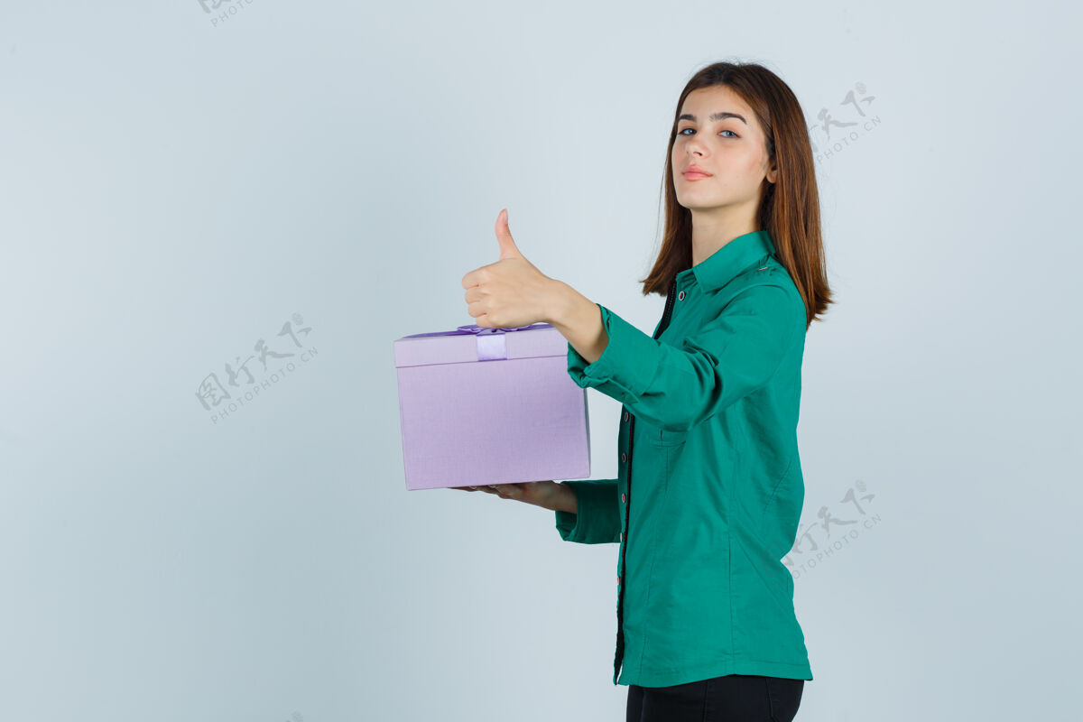 女孩拿着礼盒的年轻女孩 穿着绿色上衣 黑色裤子 竖起大拇指 看起来很自信 正面照商务专业正面