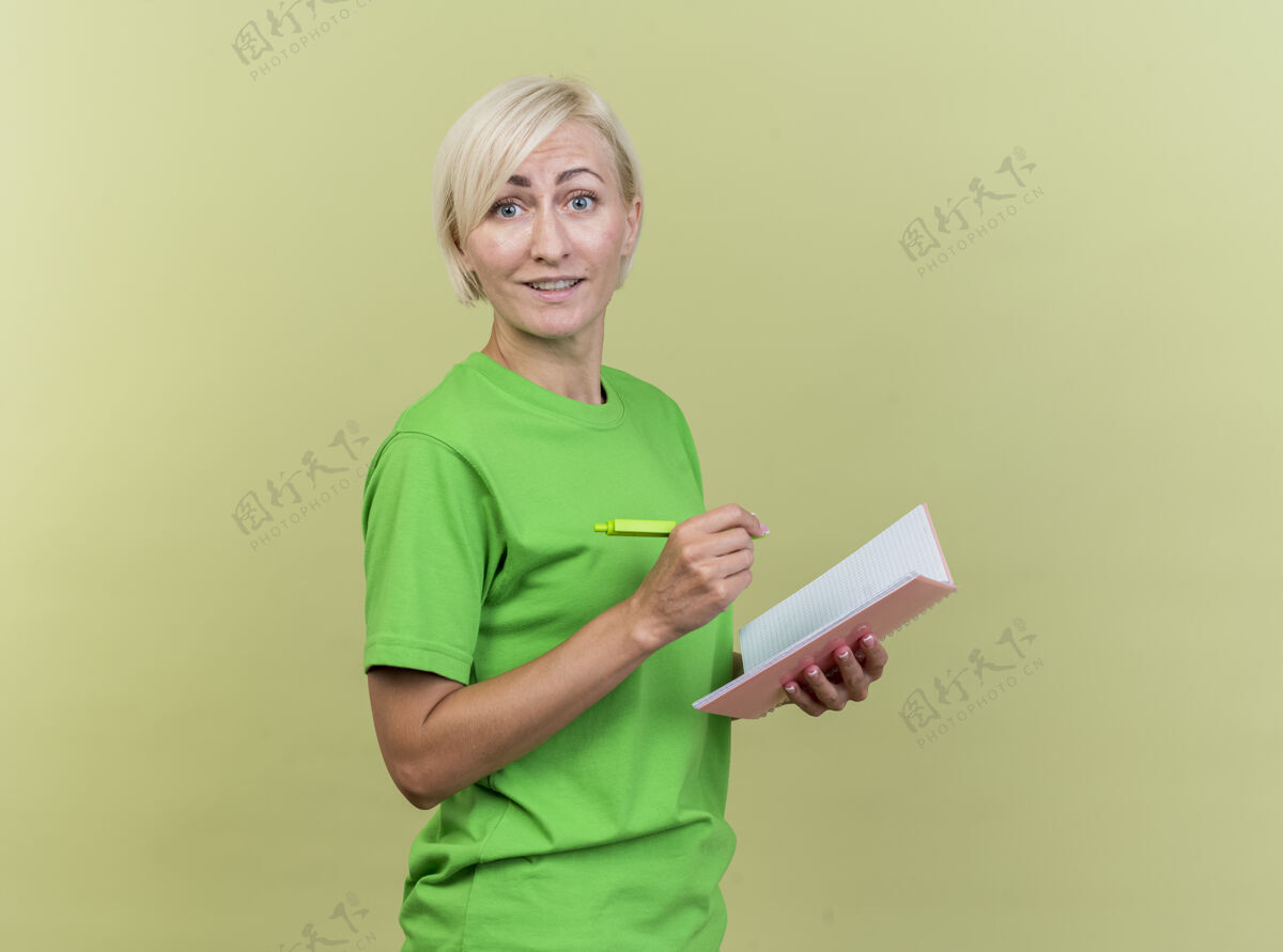 请高兴的中年金发斯拉夫妇女站在侧视图中看着相机拿着钢笔和笔记本在橄榄绿的背景与复印空间隔离视图站着复印件